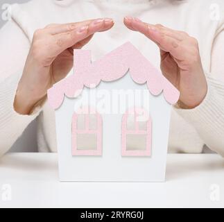 Le mani femminili si piegano l'una all'altra su una casa in miniatura in legno su sfondo bianco. Concetto di assicurazione immobiliare, envi Foto Stock