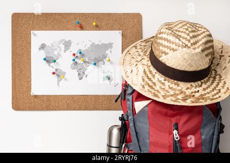 Pianificazione del concetto di destinazione di viaggio. Corkboard con mappa del mondo e spille sulla parete, zaino e cappello estivo Foto Stock