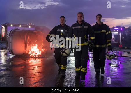 Coraggiosi pompieri che camminano verso la telecamera. In background paramedici e squadra di soccorso dei pompieri combattono Fire in Car Accident, Insu Foto Stock