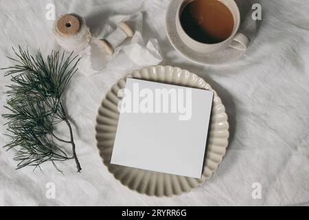 Cartoleria natalizia. Biglietto d'auguri quadrato vuoto, mockup di invito, piastra in ceramica. Nastro di seta e rami di pino. Tovaglia di lino bianca. Tazza da Foto Stock