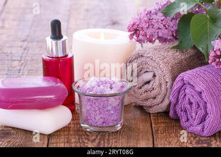 Rosso bottiglia con olio aromatico, candela che brucia, sapone, ciotola con sale marino, fiori lilla e asciugamani per il bagno le procedure su tavole di legno. Prodotto Spa Foto Stock