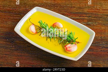 Un tavolo di legno con una ciotola bianca piena di olio d'oliva, spicchi d'aglio freschi e ciuffi di rosmarino. Foto Stock