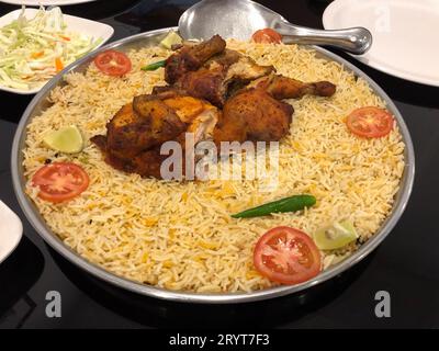 Biryani di pollo serviti su un grande piatto di acciaio e guarniti con pomodori a fette, brioche verdi e spicchi di limone. Foto Stock