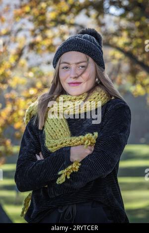 Un'immagine ravvicinata di una giovane donna che si sente fredda, vestita con un accogliente maglione nero e un cappello di lana mentre si trova sotto alberi autunnali dorati Foto Stock