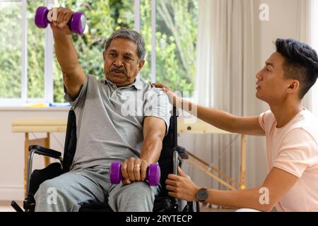 Fisioterapista maschile eterogeneo che consiglia e paziente anziano in sedia a rotelle con manubri Foto Stock