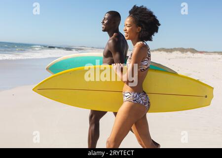 Felice coppia afroamericana in forma che porta tavole da surf a piedi sulla spiaggia soleggiata fino al mare Foto Stock