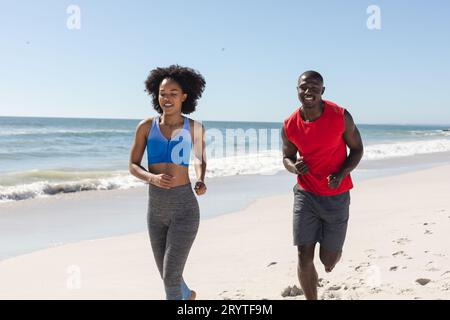 Una coppia afroamericana in forma e felice, che corre su una spiaggia soleggiata Foto Stock