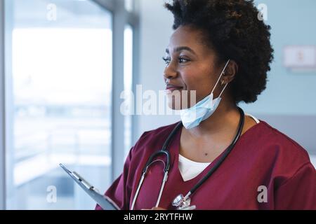Felice medico afroamericano donna che indossa scrub e maschera facciale, guardando attraverso la finestra Foto Stock