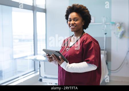 Ritratto di una felice dottoressa afroamericana che indossa scrub e stetoscopio, tenendo in mano una tavoletta Foto Stock