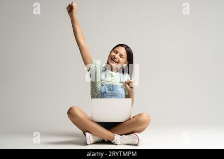Felice ragazza adolescente sorridente che rallegra seduta su sfondo grigio con il laptop sulle gambe. Foto Stock
