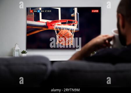 Uomo che guarda una partita di basket sulla Smart TV mentre beve un caffè. Immagine della palla che entra nel cerchio. Trasmissione TV in diretta. Foto Stock