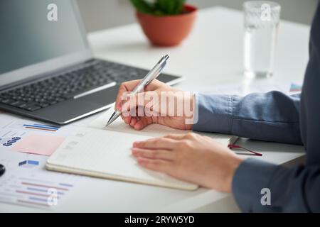 Concentratevi sulla mano di una giovane donna d'affari in camicia blu con penna su una pagina vuota del notebook durante l'organizzazione del lavoro e la stesura dell'agenda Foto Stock