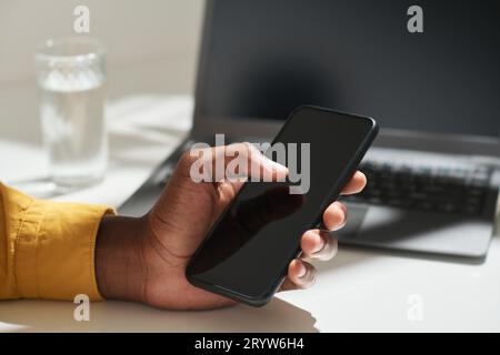 Mano di un giovane impiegato afro-americano che tocca lo schermo dello smartphone con il pollice mentre è seduto alla scrivania e sta cercando in Internet Foto Stock