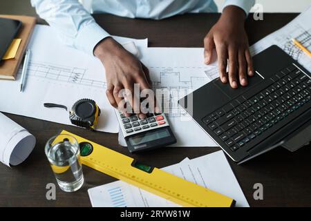 Sopra l'angolo di mano del giovane architetto afro-americano maschio con computer portatile e calcolatrice seduti sul posto di lavoro con progetti e strumenti manuali Foto Stock