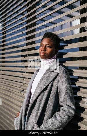 Donna afro-americana pensiva in elegante capispalla che tiene le mani in tasca e guarda lontano mentre si appoggia alle pareti contemporanee nelle giornate di sole Foto Stock