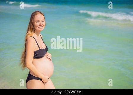 Radiosa donna incinta in costume da bagno, sullo sfondo mozzafiato di un mare turchese. Bellezza serena della maternità vicino alla riva Foto Stock