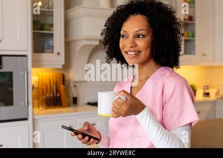 Felice lavoratrice medica birazziale in scrub bevendo caffè e utilizzando smartphone in cucina. Servizi medici, comunicazione, stile di vita e domestico Foto Stock