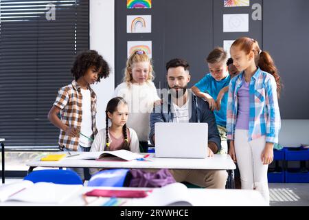 Diversi scolari elementari intorno a insegnanti maschi che utilizzano un computer portatile alla scrivania in classe, spazio per le copie Foto Stock