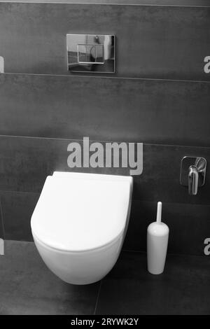 Un moderno WC bianco montato a parete con coperchio chiuso e un pulsante cromato lucido contro una parete nera. WC all'interno del bagno Foto Stock