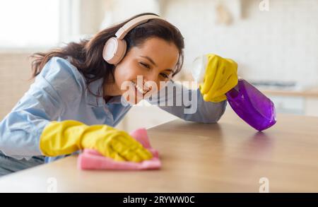 Una donna felice che spolvera il tavolo per la pulizia con lo straccio che si prende cura dei mobili in legno a casa e ascolta la musica con le cuffie Foto Stock