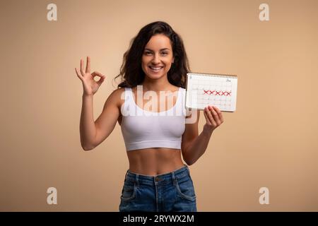 La giovane bruna felice mostra il calendario dei periodi, mostrando un gesto discreto Foto Stock