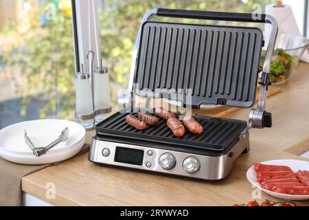 Moderna griglia elettrica con saporite salsicce su ripiano in legno in cucina Foto Stock