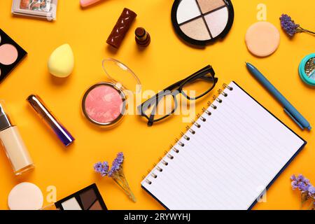 Composizione con cosmetici per il trucco, occhiali da vista, splendidi fiori e quaderno su sfondo arancione Foto Stock