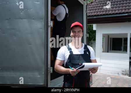 Addetto alla consegna in uniforme e berretto con leggio in piedi vicino al carrello Foto Stock