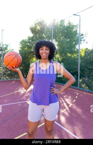 Ritratto di una felice giocatrice di basket birazziale che indossa un pallacanestro in campo da basket Foto Stock