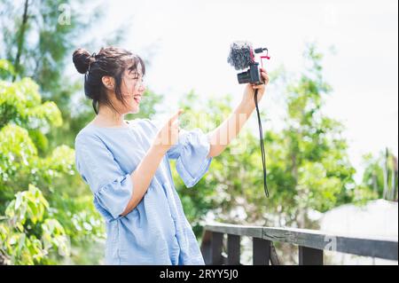 Donna asiatica in abito blu nel parco pubblico con fotocamera mirrorless digitale e foto e vlog di buon umore. La vita delle persone Foto Stock