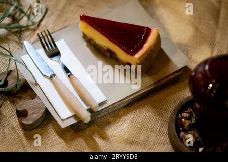 Una cattura ravvicinata di una fetta di cheesecake di fragole su una tavola rustica di legno, con il delicato abbraccio di una candela ambientale, presentata su un burlap ta Foto Stock