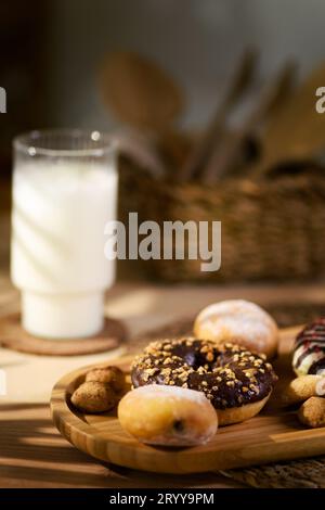 Un vassoio di legno rustico presenta tre ciambelle, biscotti ripieni di cioccolato e biscotti piccoli. Un bicchiere di latte completa la scena, insieme a un BAS Foto Stock