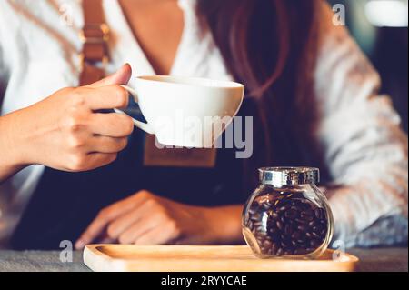 Primo piano con una barista professionista che prepara a mano e tiene in mano una tazza di caffè bianco. Giovane donna felice al bancone bar di restaura Foto Stock