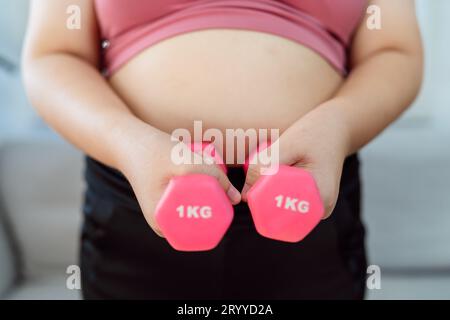 Donna in sovrappeso che si esercita per perdita di peso. Allenati con i manubri in posizioni strette a casa tua in salotto allegramente Foto Stock