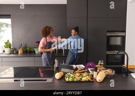 Coppia felice e variegata in grembiuli che prepara il pasto divertendosi a ballare in cucina Foto Stock