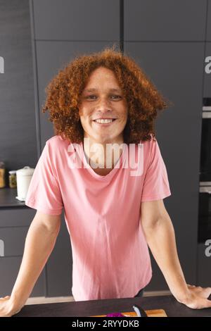 Ritratto di un felice uomo birazziale con capelli rossi ricci appoggiati al bancone nella cucina moderna Foto Stock