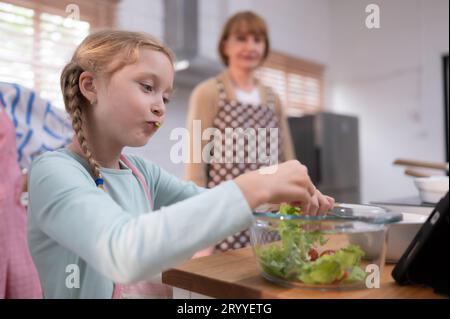 Nipote di nonni in una cucina piena di luce naturale, mangia l'insalata nonna preparata per la cena in famiglia. Foto Stock