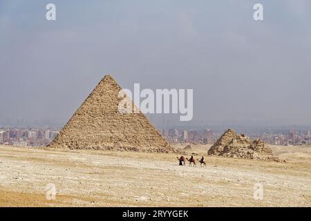 Guide a cammello e cammelli di fronte alla Piramide di Mykerinos e alle Piramidi della Regina, deserto, sabbia, calore, smog, Giza, Egitto Foto Stock