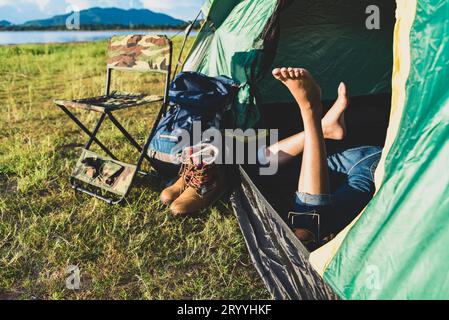 Primo piano delle gambe delle donne che si rilassano in una tenda da campeggio con lago di montagna, prato e prato sullo sfondo di prati erbosi. Stili di vita e persone Foto Stock
