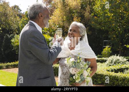 Felice sposo birazziale e sposo che beve un brindisi con champagne in giardino durante la soleggiata cerimonia nuziale Foto Stock