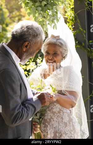 Felice sposo birazziale senior che mette un anello sul dito della sposa alla soleggiata cerimonia nuziale all'aperto Foto Stock