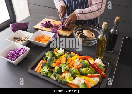 Donna birazziale anziana che indossa il grembiule e taglia il cavolo sul tavolo con verdure in cucina a casa Foto Stock