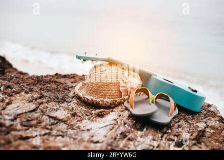 Accessori donna in vacanza in spiaggia. Ukulele, sandali e cappello sulla roccia accanto al mare. Stile tropicale e stagionale. Estate e. Foto Stock