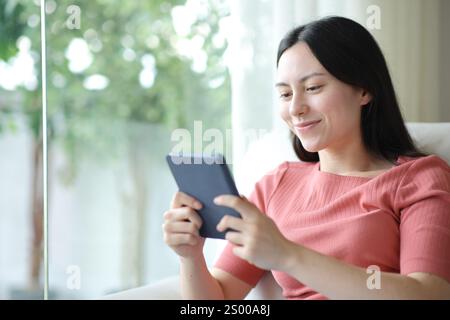 Donna asiatica soddisfatta che legge eBook a casa seduta su un divano a casa Foto Stock