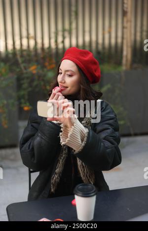 Una giovane donna elegante che indossa un vivace berretto rosso ama sorseggiare un caffè mentre si fa un divertente selfie all'aperto Foto Stock