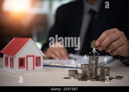 Accatastare a mano le monete vicino a un modello di casa in miniatura, simboleggiando gli investimenti immobiliari, la crescita finanziaria e il risparmio. Ideale per i settori finanziario, bancario e. Foto Stock
