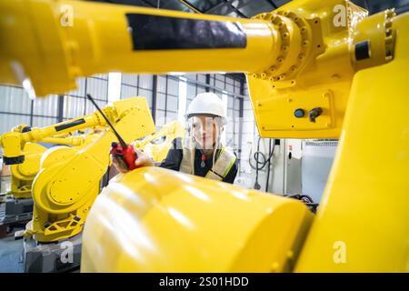 Un tecnico esperto, che indossa un elmetto e un equipaggiamento di sicurezza, lavora su bracci robotici gialli in un impianto di produzione molto affollato. L'ambiente mette in mostra l'avvento Foto Stock