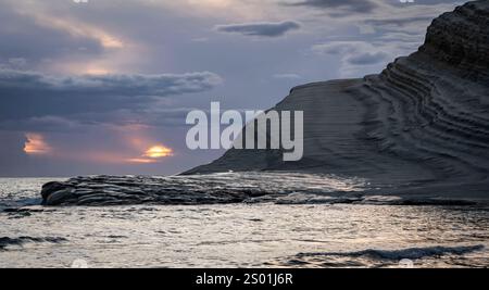 Un tranquillo paesaggio costiero caratterizzato da formazioni rocciose a strati e un mare calmo al tramonto. Il cielo è pieno di morbide nuvole che riflettono le calde tonalità dell'ora Foto Stock