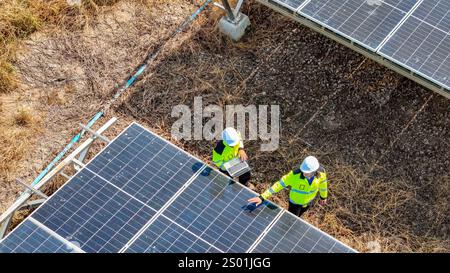 Due lavoratori che indossano cappelli rigidi e giubbotti riflettenti stanno esaminando da vicino i pannelli solari in una fattoria solare. Sono impegnati in attività di manutenzione ai sensi della lettera c Foto Stock