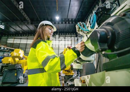 Un ingegnere esperto con giacca e elmetto giallo brillante lavora alla programmazione di apparecchiature robotiche avanzate all'interno di una struttura industriale ben illuminata. Il suo f Foto Stock